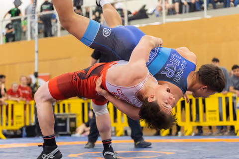 Ukrainian wrestler in blue executes a powerful throw, lifting his red-singlet opponent overhead in an indoor arena.