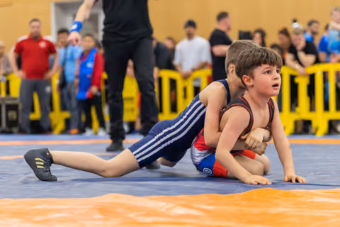 Two young boys wrestling on a blue mat, one in navy pinning the other in red, both showing intense concentration.