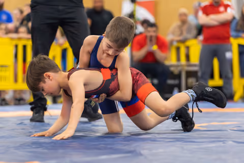 Two young boys wrestle intensely on a blue mat, one in blue dominating over the other in red, at an indoor sports event.