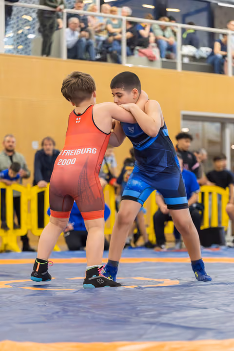 Two young wrestlers in red and blue singlets engage in a grappling match on a blue mat, with spectators watching.