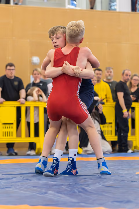 Two boys in red and blue singlets grip each other in a standing clinch, focused and determined on a blue wrestling mat.