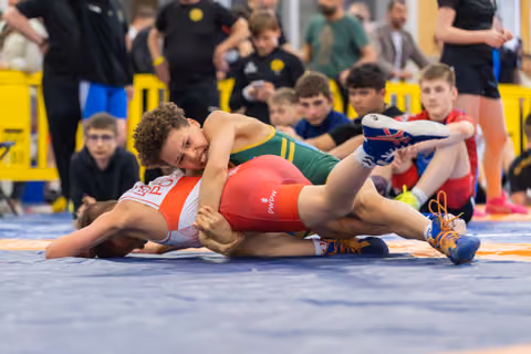 Young wrestler in green singlet pins opponent to the mat, grimacing with effort during a youth wrestling match.