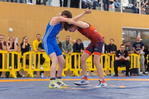 Two young wrestlers lock arms in a tense standoff on a blue mat, watched by a crowd of spectators behind yellow barriers.