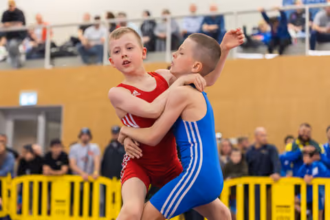 Two boys wrestle intensely in red and blue singlets, mid-grapple with determined expressions, crowd watching in background.