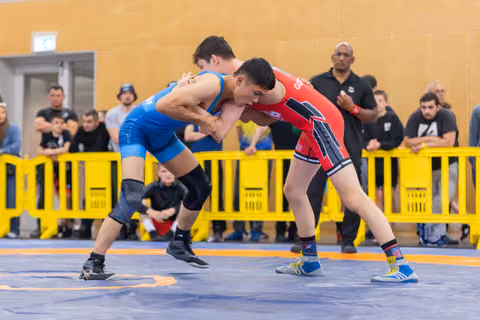 Two young wrestlers locked in intense grip, blue vs red singlet, leaning forward aggressively on competition mat with crowd watching.