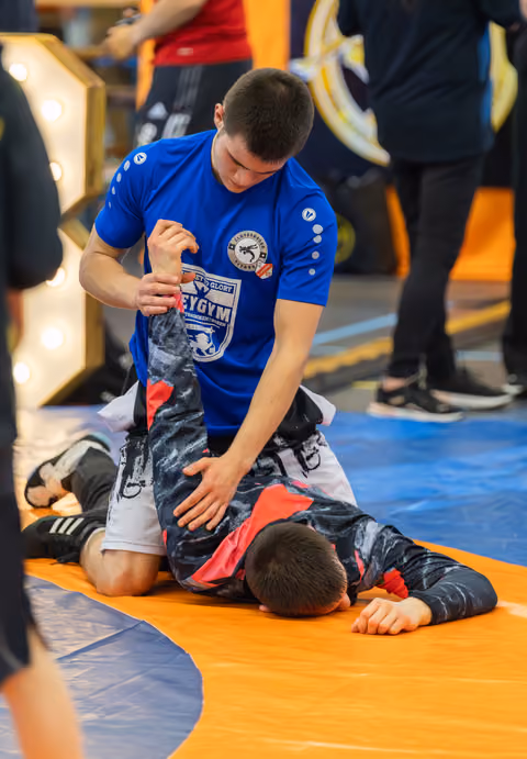 Teen in blue gym shirt applies submission hold on opponent lying face-down on orange mat during match.