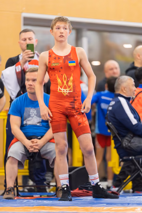 Young Ukrainian wrestler stands alert at ringside, wearing red singlet with trident emblem, composed before his match.