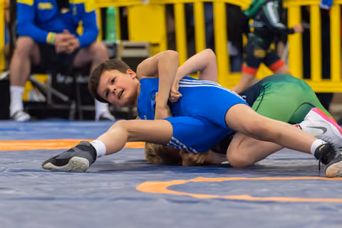 Young boy in blue singlet pins opponent to mat, face showing intense focus during youth wrestling match.