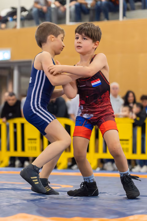Two young wrestlers grip each other's arms in a tense standoff on the mat, both focused and determined.