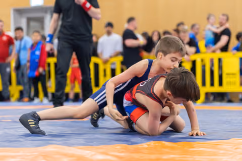 Two young wrestlers on a blue mat, one in navy pinning the other in red-black, intense focus during a youth bout.