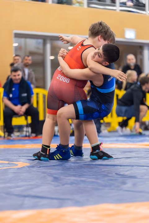 Two young wrestlers locked in a tight clinch on a blue mat, both straining with effort during a competitive bout.
