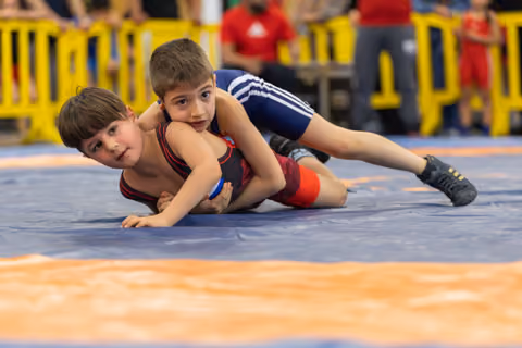 Two young boys wrestle on a blue mat, one pinning the other from behind, both showing intense focus.