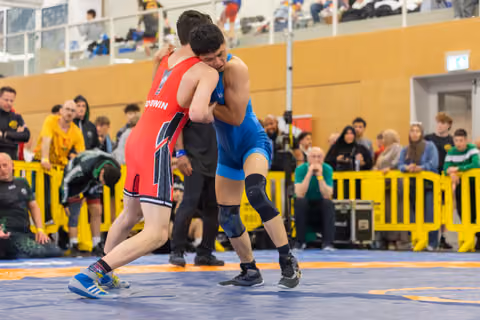 Two young wrestlers locked in a tight clinch on the mat, straining for control in front of a packed crowd.