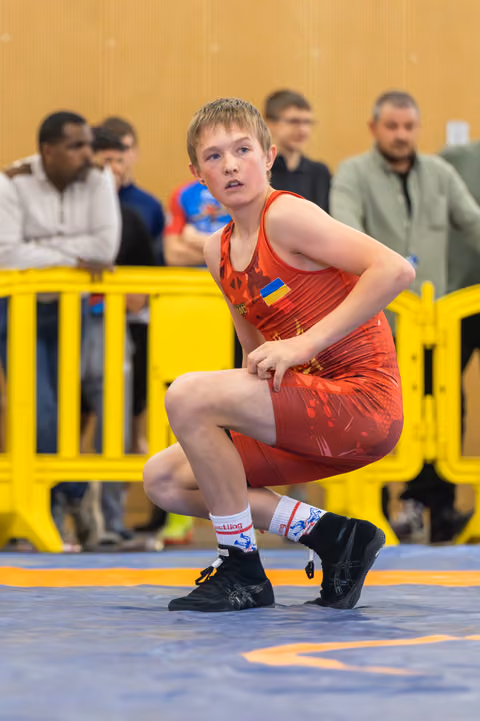 Young Ukrainian wrestler in red singlet crouches in a focused, ready stance on the blue mat during a match.