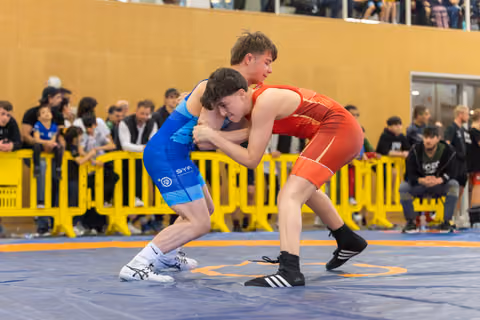 Two young wrestlers locked in a fierce clinch on the mat, spectators watching from behind yellow barriers.