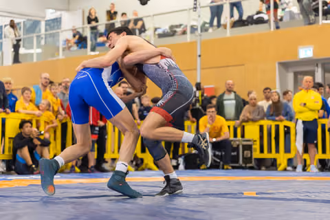 Two wrestlers locked in a tight clinch on the mat, straining against each other with focused intensity before a packed crowd.