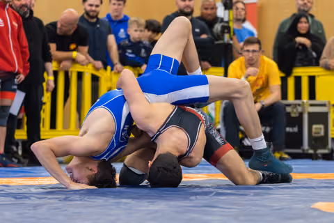 Two wrestlers grapple intensely on the mat, one in blue dominating with a takedown as spectators watch closely.