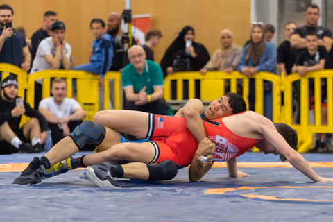 US wrestler grimaces in pain as opponent attempts a pin during an indoor match, crowd watching intently.