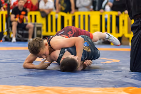 Young boy in red singlet pins his opponent to the mat, focused and determined, at an indoor youth wrestling event.
