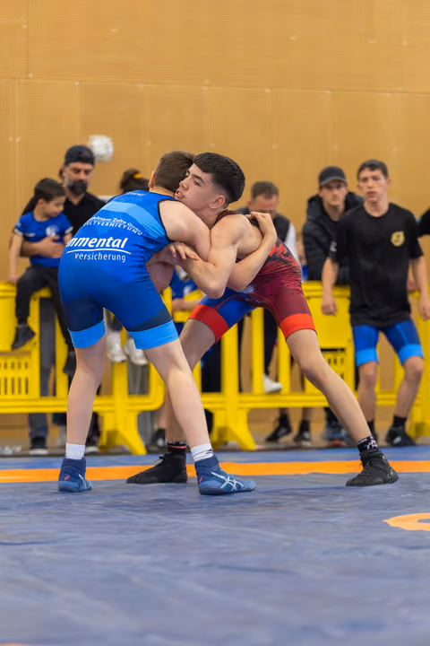 Two teenage wrestlers locked in a tight clinch on a blue mat, both straining forward with intense focus during a bout.