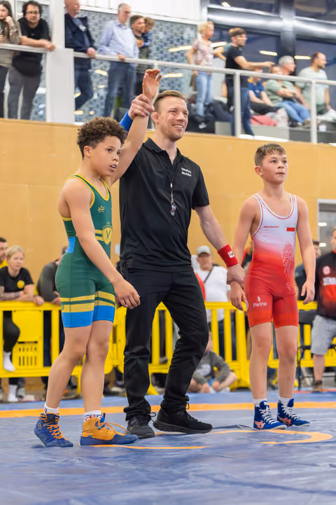 Smiling referee raises the hand of a young wrestler in green, declaring victory, as the opponent in red stands nearby.