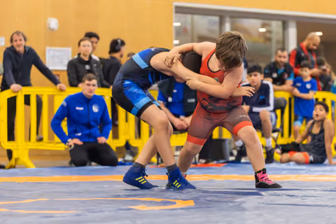 Two young wrestlers locked in a fierce clinch on the mat, leaning into each other with full effort, coaches watching nearby.
