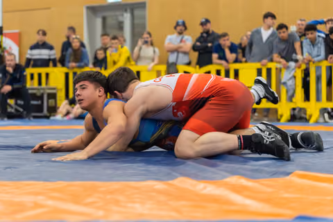 Young wrestler in blue singlet pinned down by opponent in red, struggling to escape on a blue competition mat.