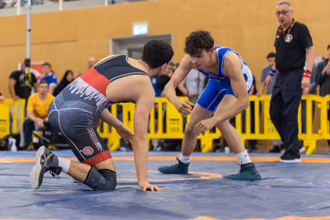 Young wrestler in blue singlet squares off aggressively over opponent on his knees on blue mat, referee watching.