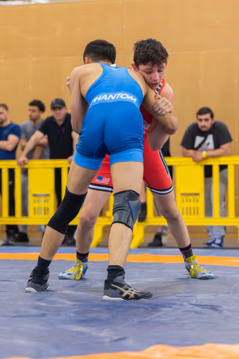 Two young wrestlers locked in a tight clinch on a blue mat, the red-singlet athlete straining with focused intensity.