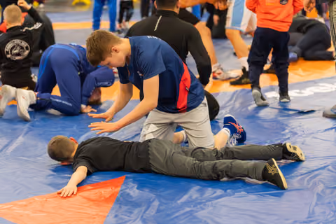 A boy in a blue shirt pins his opponent face-down on the mat, pressing firmly on his back during a match.
