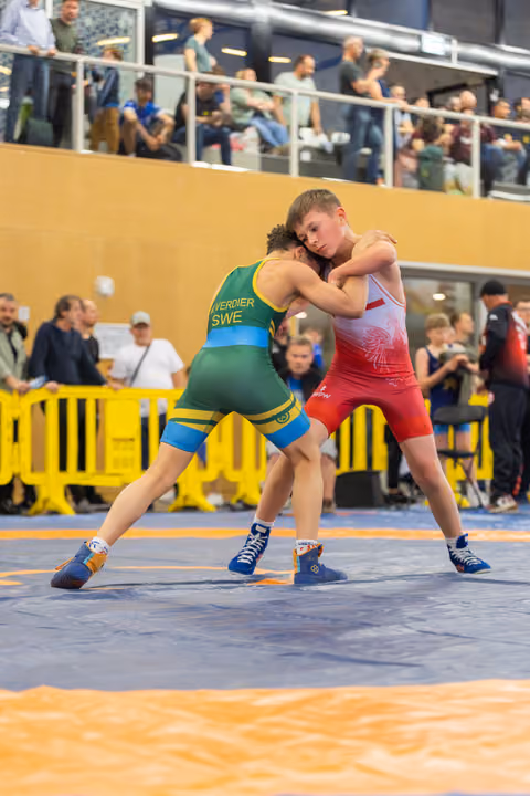 Swedish wrestler Verdier in green grapples intensely with a red-suited opponent on a blue mat before a crowded arena.