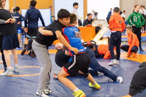 Two boys locked in an intense grapple on a blue mat, surrounded by other young wrestlers practicing in a busy hall.