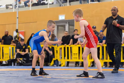 Two young wrestlers in blue and red singlets face off on a mat, referee watching closely in an indoor sports hall.