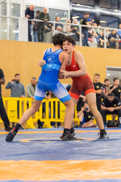 Teen in red singlet grips opponent in blue, both focused and tense on the mat, crowd watching from above.
