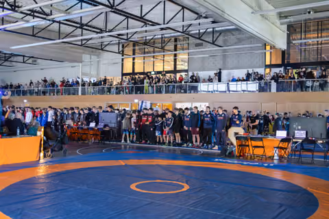 Young wrestlers stand at attention on the mat during an opening ceremony, watched by a packed crowd on the mezzanine above.