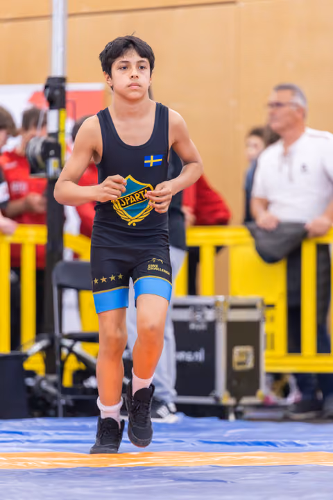 Focused young wrestler in black Sparta singlet with Swedish flag patch jogging onto the mat at an indoor sports event.