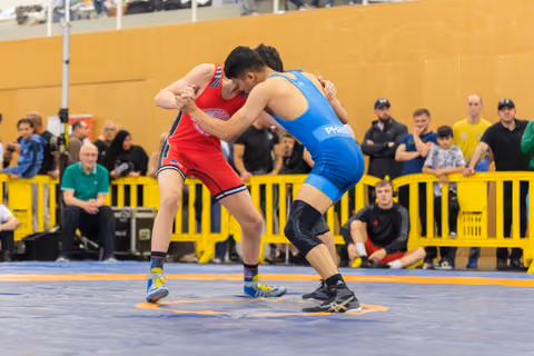 Two young wrestlers lock grips intensely on the mat, one in red, one in blue, surrounded by spectators.
