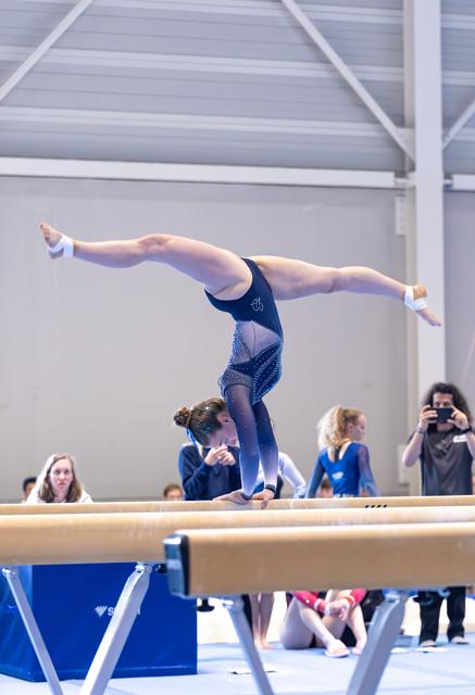 Young athlete performs a one-armed handstand on balance beam, legs split gracefully, while spectators watch in indoor venue