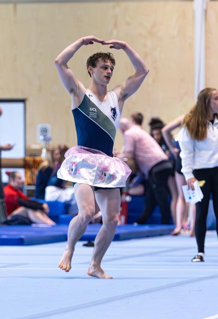 Young man performs expressive dance routine with heart-shaped hand gesture above head, wearing colorful tutu on floor exercise mat