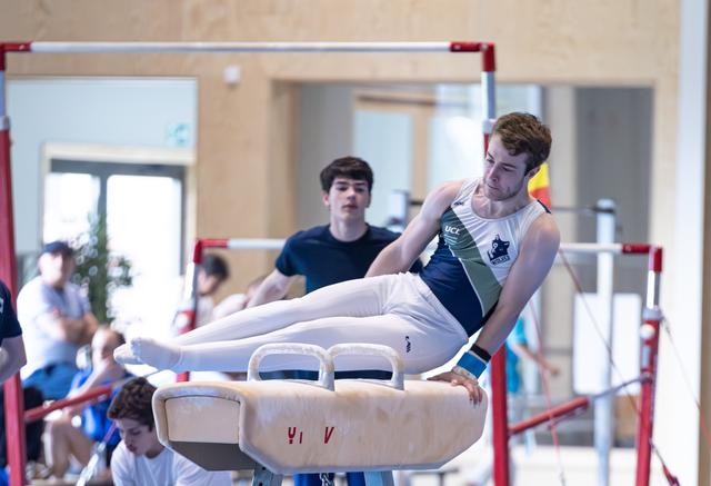 Male athlete performing pommel horse routine with extended legs while coach observes in indoor training facility